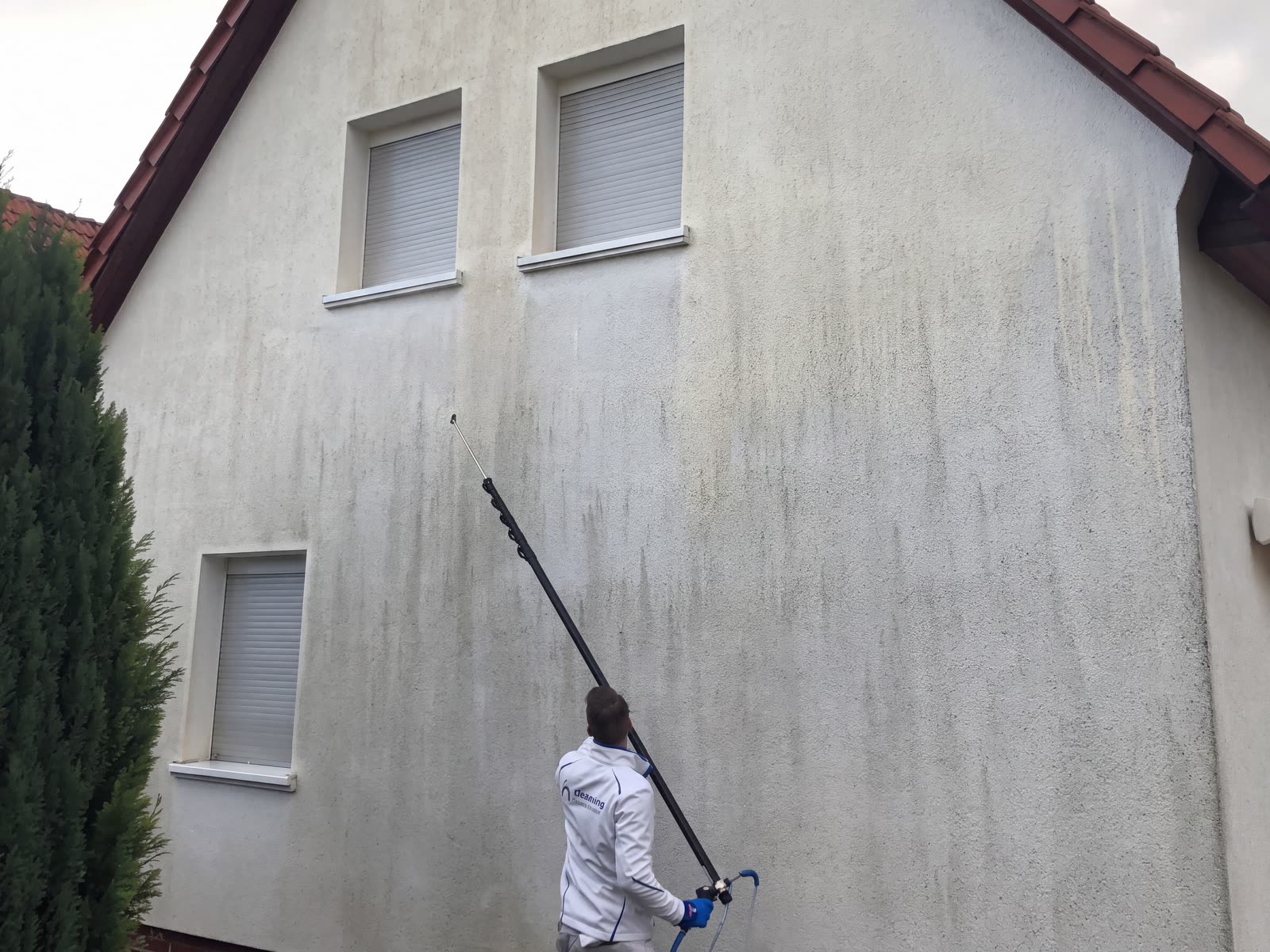 Mitarbeiter reinigt eine hohe Hauswand mit einer Teleskoplanze vom Boden aus, deutlich sichtbare saubere Bahnen.